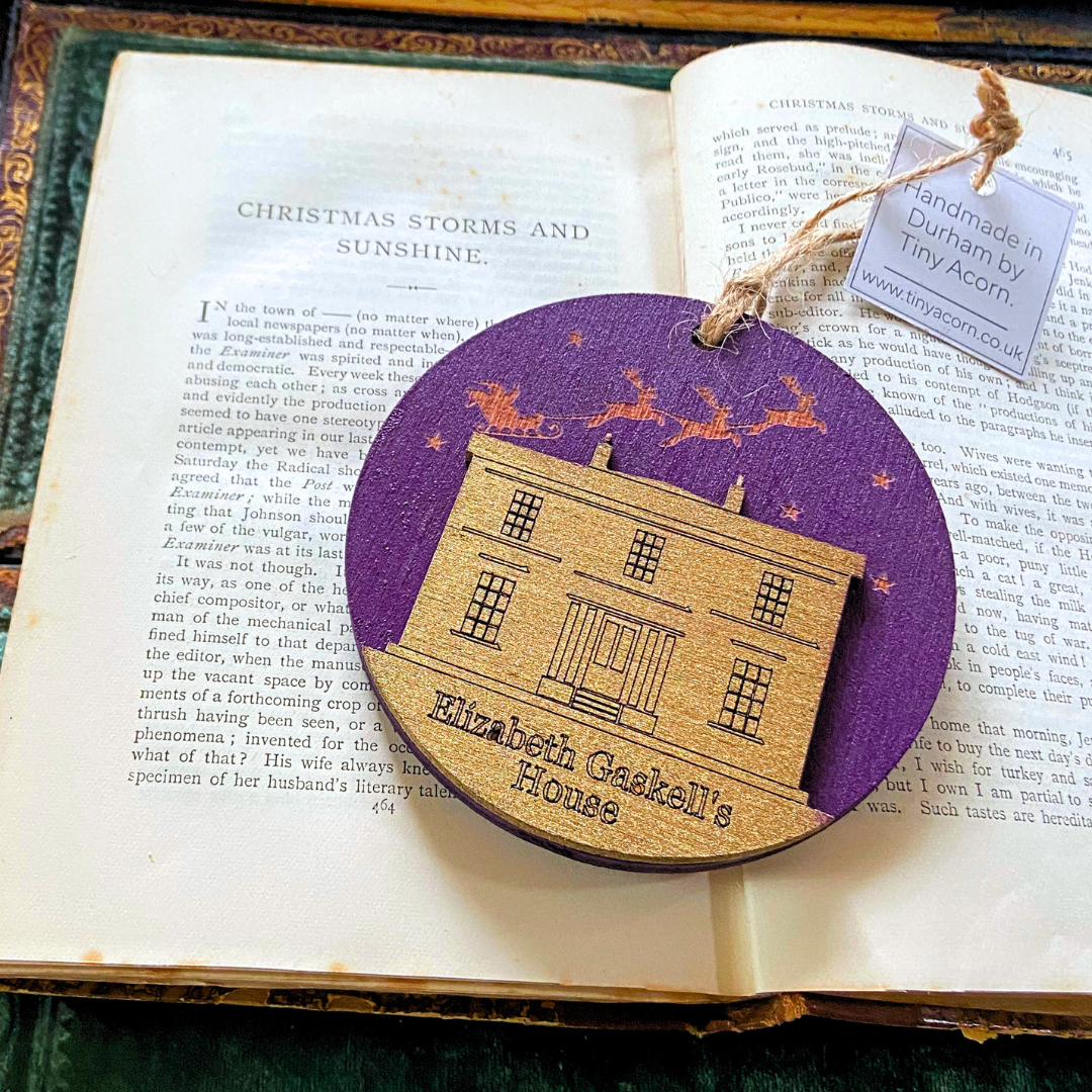 Christmas bauble featuring a golden image of Elizabeth Gaskell's House on a purple background. A gold sleigh with reindeer flies overhead. The bauble is flat and round, made of wood.