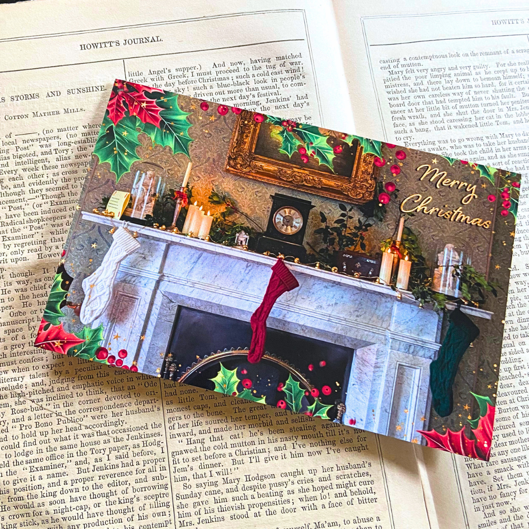An Elizabeth Gaskell's House Christmas card set against a Victorian book. The card is landscape and depicts a Victorian fireplace decorated for Christmas, with stockings and holly. It has a holly border and a Merry Christmas text graphic.